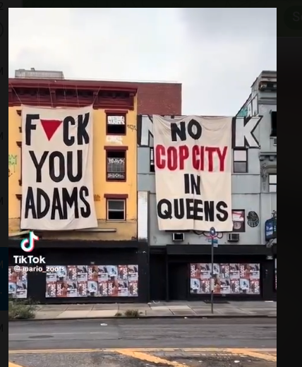 Two banners reading "Fuck you Adams" where the u in fuck is a downwards-facing red triangle and No Cop City in Queens respectively, hung from buildings.